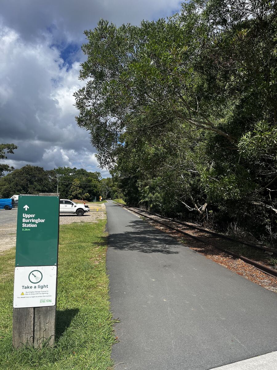 The sealed rail trail curving through trees near Upper Burringbar, with a green trail marker post